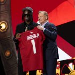 FootballR - NFL - Zwei Männer auf einer Bühne, einer jung in einer bestickten Jacke und Mütze, lächelnd, während er ein rotes Footballtrikot mit der Aufschrift Marvin Harrison Jr. 1 aus dem NFL Draft 2024 hält. Diese Beschreibung wurde automatisch generiert.
