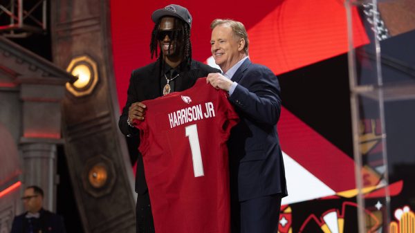 FootballR - NFL - Zwei Männer auf einer Bühne, einer jung in einer bestickten Jacke und Mütze, lächelnd, während er ein rotes Footballtrikot mit der Aufschrift Marvin Harrison Jr. 1 aus dem NFL Draft 2024 hält. Diese Beschreibung wurde automatisch generiert.