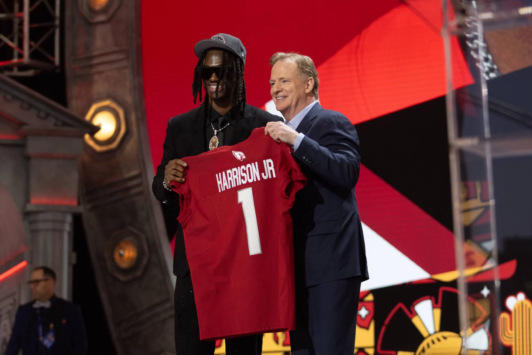 FootballR - NFL - Zwei Männer auf einer Bühne, einer jung in einer bestickten Jacke und Mütze, lächelnd, während er ein rotes Footballtrikot mit der Aufschrift Marvin Harrison Jr. 1 aus dem NFL Draft 2024 hält. Diese Beschreibung wurde automatisch generiert.