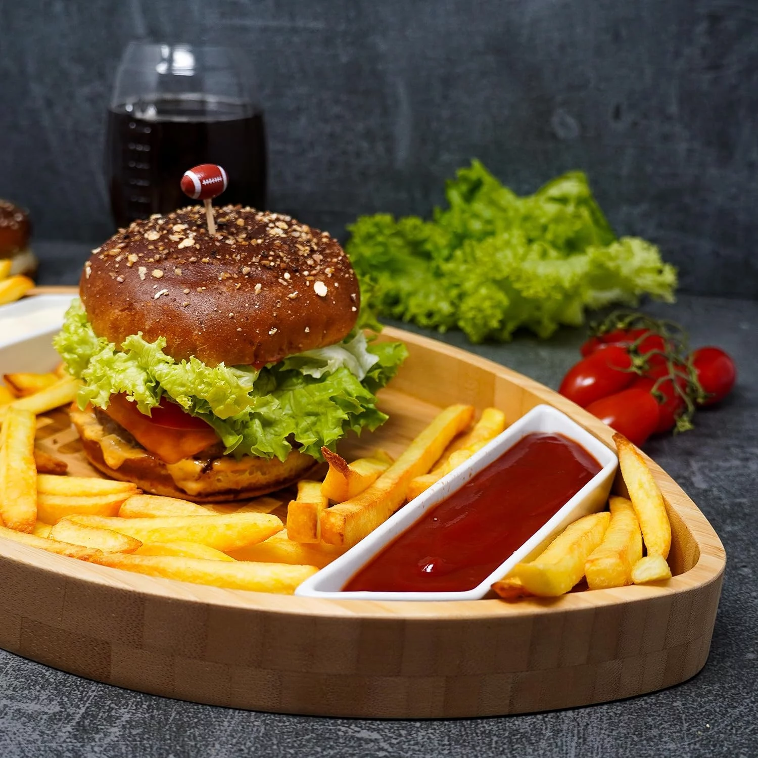 FootballR - NFL - Ein Burger mit Salat, Tomaten und einem Sesambrötchen wird mit Pommes frites und einer rechteckigen Schale Ketchup auf einer Holzplatte serviert. Ein kleiner Fußballzahnstocher ziert den Burger und deutet auf Ihre Liebe zum Spiel hin. Im Hintergrund liegen frischer Salat und Kirschtomaten neben einem NFL-Bettwäscheset auf einer dunklen Oberfläche. Diese Beschreibung wurde automatisch generiert.