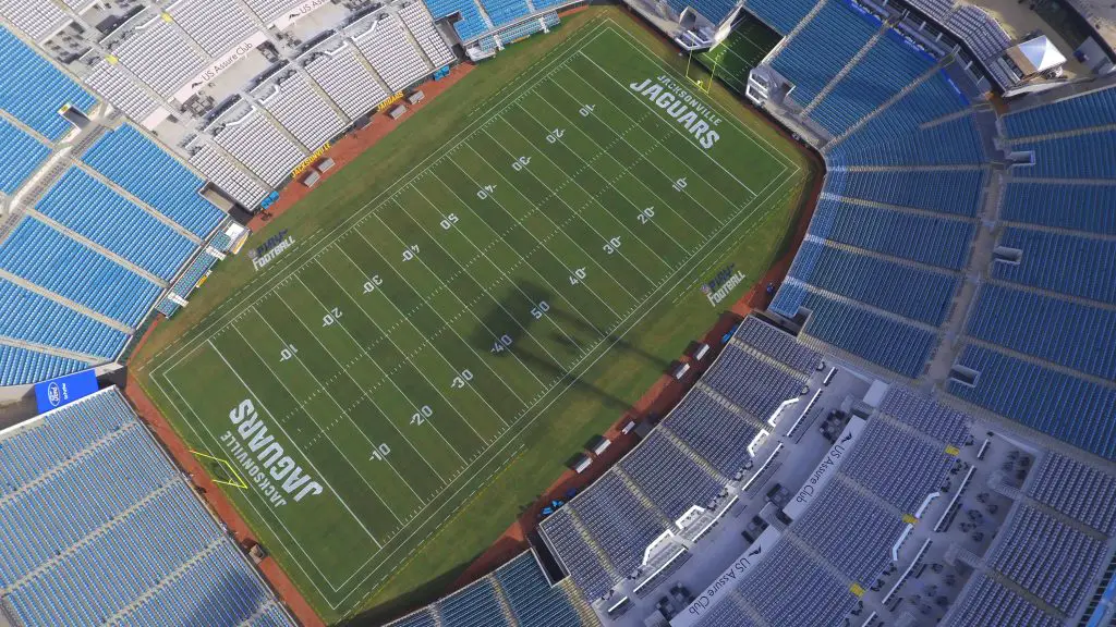 FootballR - NFL Jacksonville Jaguars - Luftaufnahme eines großen, leeren Footballstadions mit blauen Sitzen. Das Spielfeld, das mit weißen Yard-Linien und „JAGUARS“ in den Endzonen markiert ist, gehört den Jacksonville Jaguars. Imbissstände und andere Strukturen sind rund um den Sitzbereich sichtbar. Die Beleuchtung des Stadions wirft einen Schatten auf das Spielfeld. Diese Beschreibung wurde automatisch generiert.