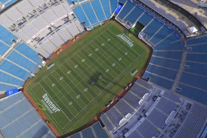 FootballR - NFL Jacksonville Jaguars - Luftaufnahme eines großen, leeren Footballstadions mit blauen Sitzen. Das Spielfeld, das mit weißen Yard-Linien und „JAGUARS“ in den Endzonen markiert ist, gehört den Jacksonville Jaguars. Imbissstände und andere Strukturen sind rund um den Sitzbereich sichtbar. Die Beleuchtung des Stadions wirft einen Schatten auf das Spielfeld. Diese Beschreibung wurde automatisch generiert.