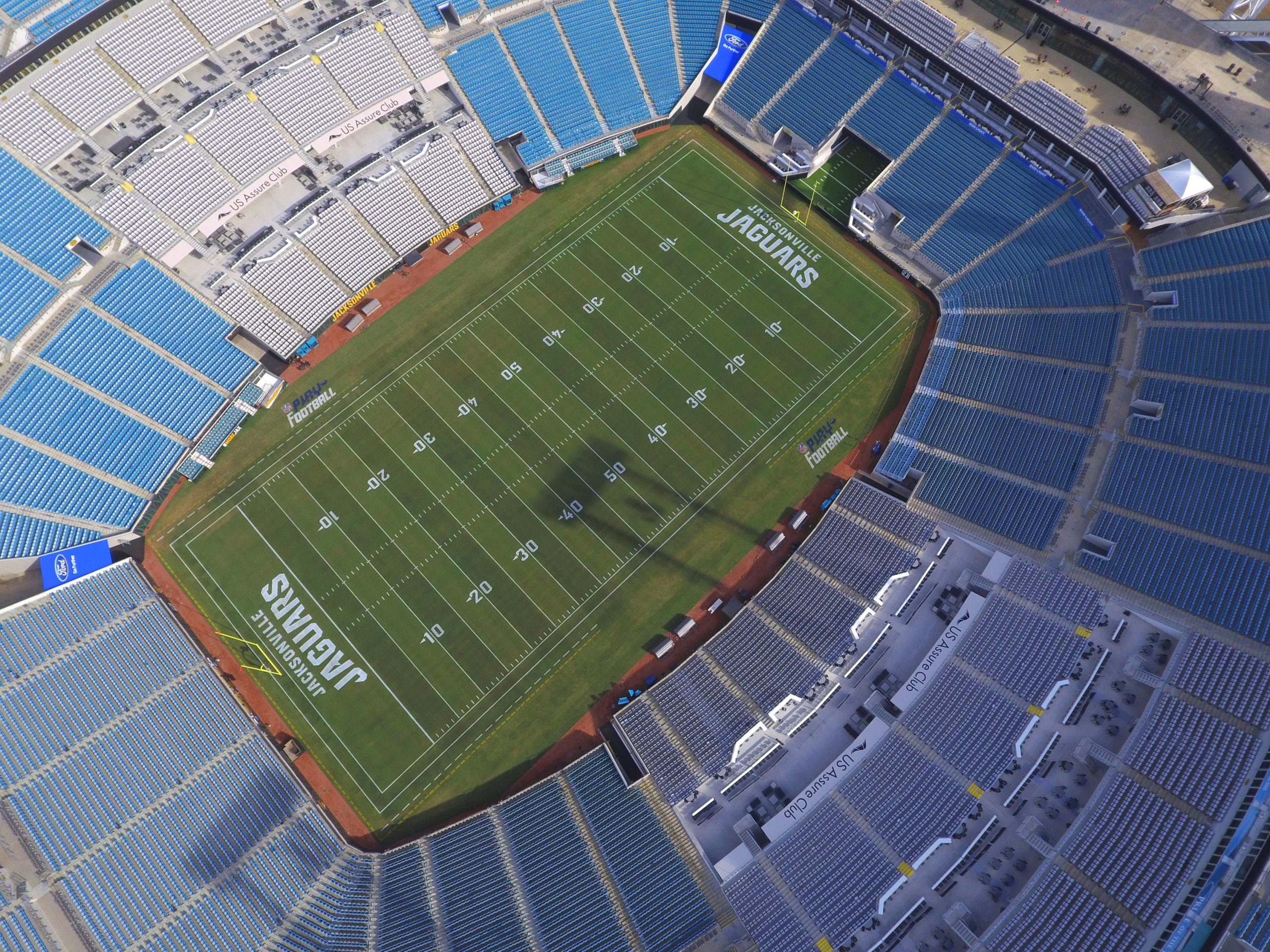 FootballR - NFL Jacksonville Jaguars - Luftaufnahme eines großen, leeren Footballstadions mit blauen Sitzen. Das Spielfeld, das mit weißen Yard-Linien und „JAGUARS“ in den Endzonen markiert ist, gehört den Jacksonville Jaguars. Imbissstände und andere Strukturen sind rund um den Sitzbereich sichtbar. Die Beleuchtung des Stadions wirft einen Schatten auf das Spielfeld. Diese Beschreibung wurde automatisch generiert.
