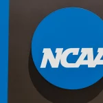FootballR - NFL - Das Bild zeigt das NCAA-Logo mit dem Akronym „NCAA“ in fetten weißen Buchstaben in der Mitte eines blauen Kreises. Der blaue Kreis befindet sich auf einem grauen Hintergrund mit einem vertikalen blauen Streifen auf der linken Seite des Bildes, der die Traditionen des Hochschulsports und Diskussionen rund um die Spielerbezahlung symbolisiert. Diese Beschreibung wurde automatisch generiert.