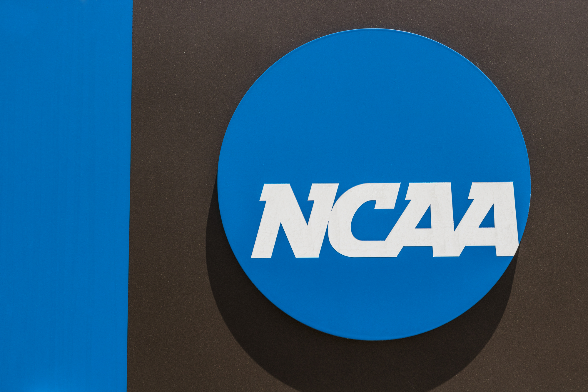 FootballR - NFL - Das Bild zeigt das NCAA-Logo mit dem Akronym „NCAA“ in fetten weißen Buchstaben in der Mitte eines blauen Kreises. Der blaue Kreis befindet sich auf einem grauen Hintergrund mit einem vertikalen blauen Streifen auf der linken Seite des Bildes, der die Traditionen des Hochschulsports und Diskussionen rund um die Spielerbezahlung symbolisiert. Diese Beschreibung wurde automatisch generiert.