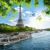 FootballR - NFL Paris und Dublin - Eine malerische Aussicht auf den Eiffelturm in Paris, Frankreich, unter einem strahlend blauen Himmel mit vereinzelten Wolken. Im Vordergrund fließt die Seine, auf der ein Ausflugsboot kreuzt. Grüne Blätter rahmen den oberen und seitlichen Teil des Bildes ein und verleihen dieser ikonischen Pariser Szene einen lebendigen Hauch von Natur. Diese Beschreibung wurde automatisch generiert.