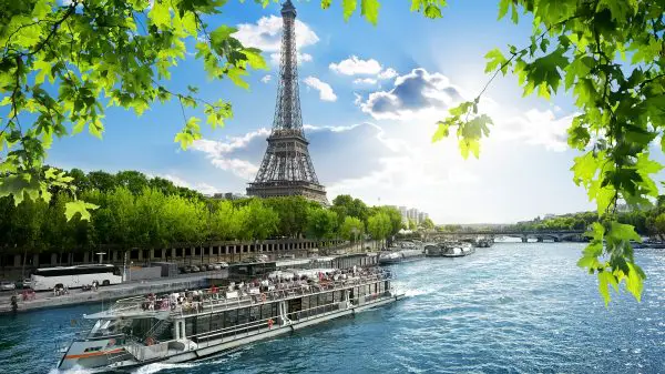 FootballR - NFL Paris und Dublin - Eine malerische Aussicht auf den Eiffelturm in Paris, Frankreich, unter einem strahlend blauen Himmel mit vereinzelten Wolken. Im Vordergrund fließt die Seine, auf der ein Ausflugsboot kreuzt. Grüne Blätter rahmen den oberen und seitlichen Teil des Bildes ein und verleihen dieser ikonischen Pariser Szene einen lebendigen Hauch von Natur. Diese Beschreibung wurde automatisch generiert.