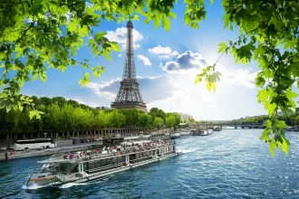 FootballR - NFL Paris und Dublin - Eine malerische Aussicht auf den Eiffelturm in Paris, Frankreich, unter einem strahlend blauen Himmel mit vereinzelten Wolken. Im Vordergrund fließt die Seine, auf der ein Ausflugsboot kreuzt. Grüne Blätter rahmen den oberen und seitlichen Teil des Bildes ein und verleihen dieser ikonischen Pariser Szene einen lebendigen Hauch von Natur. Diese Beschreibung wurde automatisch generiert.