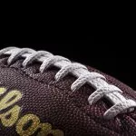 Championship Game - Prime Days - NFL - Nahaufnahme eines traditionellen American Footballs vor schwarzem Hintergrund. Die strukturierte Oberfläche aus braunem Leder ist sichtbar, mit weißen Schnürsenkeln in der Mitte. Die Worte „Wilson NFL Duke Replica“ sind in Gold auf den Ball gedruckt und geben die Marke und das Modell an. Der Fokus hebt die Nähte und die Maserung hervor. Diese Beschreibung wurde automatisch generiert. Nordic Storm