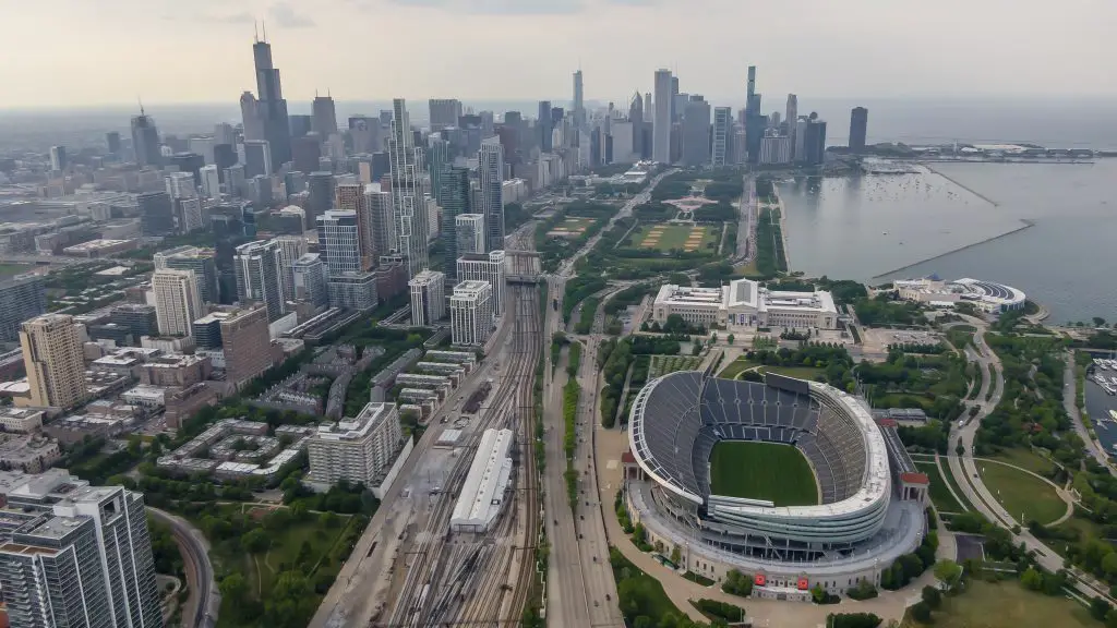 FootballR - NFL - Luftaufnahme von Chicago mit der Skyline mit hoch aufragenden Wolkenkratzern, dem Michigansee, Soldier Field, der Heimat der Chicago Bears, und mehreren zusammenlaufenden Bahngleisen an einem bewölkten Tag. Diese Beschreibung wurde automatisch generiert.