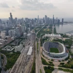 FootballR - NFL - Luftaufnahme von Chicago mit der Skyline mit hoch aufragenden Wolkenkratzern, dem Michigansee, Soldier Field, der Heimat der Chicago Bears, und mehreren zusammenlaufenden Bahngleisen an einem bewölkten Tag. Diese Beschreibung wurde automatisch generiert.