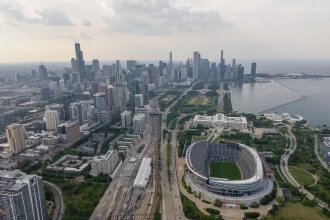FootballR - NFL - Luftaufnahme von Chicago mit der Skyline mit hoch aufragenden Wolkenkratzern, dem Michigansee, Soldier Field, der Heimat der Chicago Bears, und mehreren zusammenlaufenden Bahngleisen an einem bewölkten Tag. Diese Beschreibung wurde automatisch generiert.