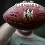 FootballR - NFL - Nürnberg Rams - NFLPA - Eine Person hält einen offiziellen NFL-Football in die Kamera und verdeckt dabei ihr Gesicht. Auf dem Football sind das NFL-Logo, „The Duke“, das Wilson-Branding und der Schriftzug „Commissioner Roger Goodell“ zu sehen. Der Hintergrund ist dunkel und verschwommen, während die Person ein kurzärmeliges Hemd trägt, das wahrscheinlich außerhalb der Saison aufgenommen wurde. Diese Beschreibung wurde automatisch generiert.