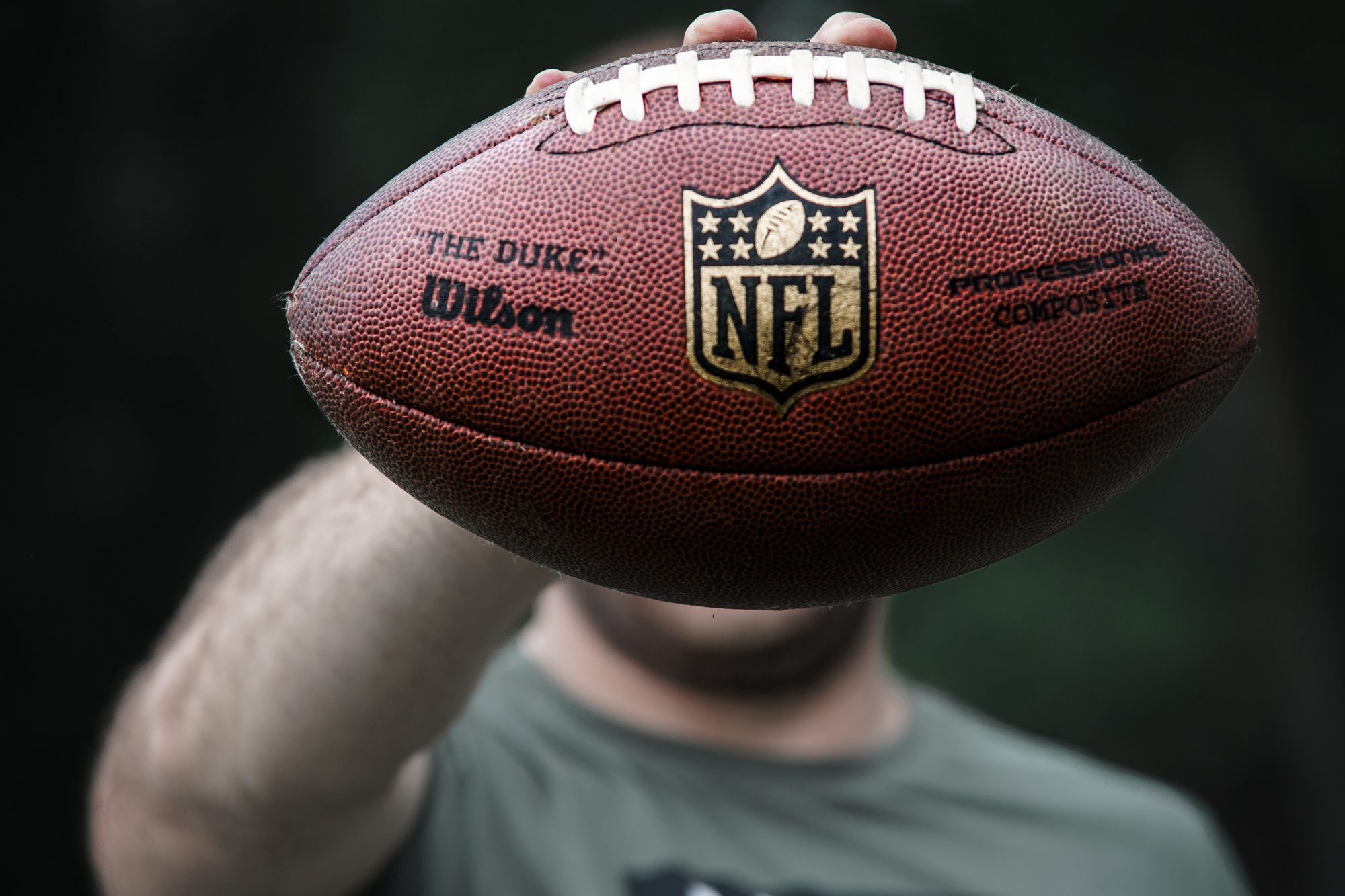 FootballR - NFL - Nürnberg Rams - NFLPA - Eine Person hält einen offiziellen NFL-Football in die Kamera und verdeckt dabei ihr Gesicht. Auf dem Football sind das NFL-Logo, „The Duke“, das Wilson-Branding und der Schriftzug „Commissioner Roger Goodell“ zu sehen. Der Hintergrund ist dunkel und verschwommen, während die Person ein kurzärmeliges Hemd trägt, das wahrscheinlich außerhalb der Saison aufgenommen wurde. Diese Beschreibung wurde automatisch generiert.