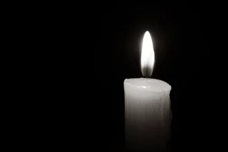 Jeremiah Kelly - NFL - Mason Alexander - Eine einzelne weiße Kerze, an deren Seite geschmolzenes Wachs heruntertropft, brennt hell vor einem pechschwarzen Hintergrund. Die Flamme ist gleichmäßig und leuchtend, wirft einen sanften Schein um die Kerze und beleuchtet ihre wachsartige Oberfläche. Der starke Kontrast, ähnlich wie Jim Ottos Vermächtnis als Mr. Raider, betont das zarte Licht der Kerze. Diese Beschreibung wurde automatisch generiert.