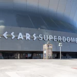 FootballR - NFL - Weitwinkelansicht des Äußeren des Caesars Superdome an einem sonnigen Tag, die seine große, geschwungene Metallfassade zeigt. Der Name des Veranstaltungsortes, „Caesars Superdome“, ist in großen, fetten Buchstaben deutlich zu sehen. Unter der Hauptfassade sind einige kleinere Eingänge und Schilder zu sehen. Der umliegende Bereich ist geräumig und leer, bereit, die Fans der New Orleans Saints willkommen zu heißen. Diese Beschreibung wurde automatisch generiert.