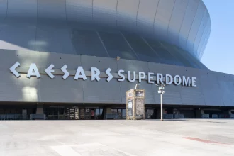 FootballR - Domecoming - Weitwinkelansicht des Äußeren des Caesars Superdome an einem sonnigen Tag, die seine große, geschwungene Metallfassade zeigt. Der Name des Veranstaltungsortes, „Caesars Superdome“, ist in großen, fetten Buchstaben deutlich zu sehen. Unter der Hauptfassade sind einige kleinere Eingänge und Schilder zu sehen. Der umliegende Bereich ist geräumig und leer, bereit, die Fans der New Orleans Saints willkommen zu heißen. Diese Beschreibung wurde automatisch generiert.