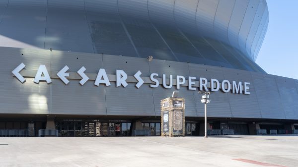 FootballR - NFL - Weitwinkelansicht des Äußeren des Caesars Superdome an einem sonnigen Tag, die seine große, geschwungene Metallfassade zeigt. Der Name des Veranstaltungsortes, „Caesars Superdome“, ist in großen, fetten Buchstaben deutlich zu sehen. Unter der Hauptfassade sind einige kleinere Eingänge und Schilder zu sehen. Der umliegende Bereich ist geräumig und leer, bereit, die Fans der New Orleans Saints willkommen zu heißen. Diese Beschreibung wurde automatisch generiert.