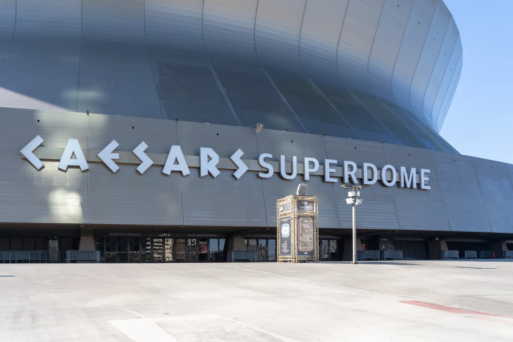 FootballR - NFL - Weitwinkelansicht des Äußeren des Caesars Superdome an einem sonnigen Tag, die seine große, geschwungene Metallfassade zeigt. Der Name des Veranstaltungsortes, „Caesars Superdome“, ist in großen, fetten Buchstaben deutlich zu sehen. Unter der Hauptfassade sind einige kleinere Eingänge und Schilder zu sehen. Der umliegende Bereich ist geräumig und leer, bereit, die Fans der New Orleans Saints willkommen zu heißen. Diese Beschreibung wurde automatisch generiert.