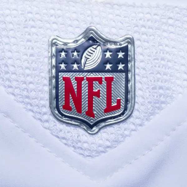 FootballR - NFL - Nahaufnahme des NFL Spielplan 2024 -Logos auf einem weißen Footballtrikot. Das Logo zeigt ein Muster der amerikanischen Flagge mit Sternen und Streifen, ein Football-Symbol und fettgedruckte rote „NFL“-Buchstaben vor Diese Beschreibung wurde automatisch generiert.