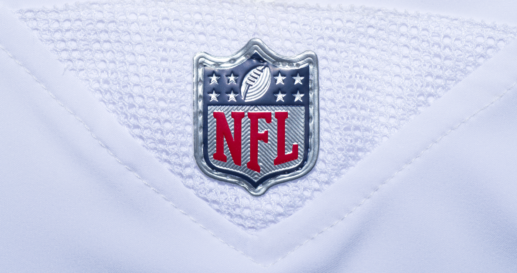 FootballR - NFL - Nahaufnahme des NFL Spielplan 2024 -Logos auf einem weißen Footballtrikot. Das Logo zeigt ein Muster der amerikanischen Flagge mit Sternen und Streifen, ein Football-Symbol und fettgedruckte rote „NFL“-Buchstaben vor Diese Beschreibung wurde automatisch generiert.