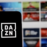 FootballR - NFL - Ein Smartphone mit dem DAZN-Logo auf dem Bildschirm wird vor einen unscharfen Hintergrund gehalten. Der Hintergrund scheint ein Computer- oder Fernsehbildschirm zu sein, auf dem verschiedene sportbezogene Miniaturansichten angezeigt werden, darunter NFL Game Pass Inhalte, wodurch der digitale Streaming-Kontext im Zusammenhang mit DAZN hervorgehoben wird. Diese Beschreibung wurde automatisch generiert.
