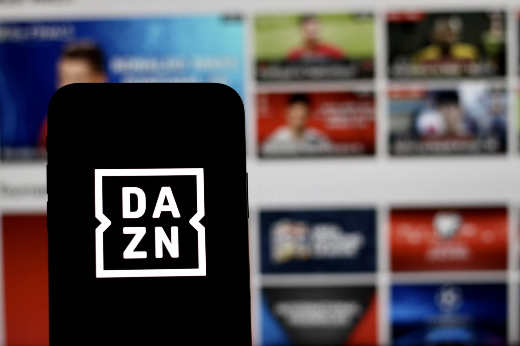 FootballR - NFL - Ein Smartphone mit dem DAZN-Logo auf dem Bildschirm wird vor einen unscharfen Hintergrund gehalten. Der Hintergrund scheint ein Computer- oder Fernsehbildschirm zu sein, auf dem verschiedene sportbezogene Miniaturansichten angezeigt werden, darunter NFL Game Pass Inhalte, wodurch der digitale Streaming-Kontext im Zusammenhang mit DAZN hervorgehoben wird. Diese Beschreibung wurde automatisch generiert.
