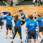 FootballR - NFL - Eine Gruppe junger Jungs in blauen Hemden feiert freudig auf einem Hallenplatz, während einer mit einem Fußball in der Hand in die Luft springt. Sie tragen passende Trikots mit dem Aufdruck „FLAG“ auf dem Rücken und schwarze Shorts mit daran befestigten blauen Flaggen. Zuschauer und Schiedsrichter in schwarz-weißen Streifen verfolgen die Aufregung der Flag-Football-Saison 2024. Diese Beschreibung wurde automatisch generiert.