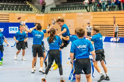 FootballR - NFL - Eine Gruppe junger Jungs in blauen Hemden feiert freudig auf einem Hallenplatz, während einer mit einem Fußball in der Hand in die Luft springt. Sie tragen passende Trikots mit dem Aufdruck „FLAG“ auf dem Rücken und schwarze Shorts mit daran befestigten blauen Flaggen. Zuschauer und Schiedsrichter in schwarz-weißen Streifen verfolgen die Aufregung der Flag-Football-Saison 2024. Diese Beschreibung wurde automatisch generiert.