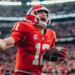 FootballR - NFL - Ein College-Footballspieler in orangefarbener Uniform, Helm und Handschuhen mit der Nummer 12 feiert einen erfolgreichen Spielzug, während er den Football hält. Man sieht den Spieler in einem voll besetzten Stadion vor Begeisterung schreien und damit zeigen, warum American Football in Europa so beliebt ist. Auf der Uniform ist ein SEC-Logo zu sehen. Diese Beschreibung wurde automatisch generiert.