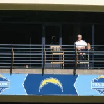 Aufzug Chargers - FootballR - NFL A.J. Smith - Ein Mann mit Sonnenbrille und weißem Poloshirt sitzt hinter einem Geländer mit dem Logo des NFL-Teams der Chargers. Er wirkt entspannt und beobachtet das Geschehen an einem sonnigen Tag von einem schattigen Balkon aus. Im Hintergrund mischen sich die General Manager verschiedener NFL-Teams untereinander. Diese Beschreibung wurde automatisch generiert.