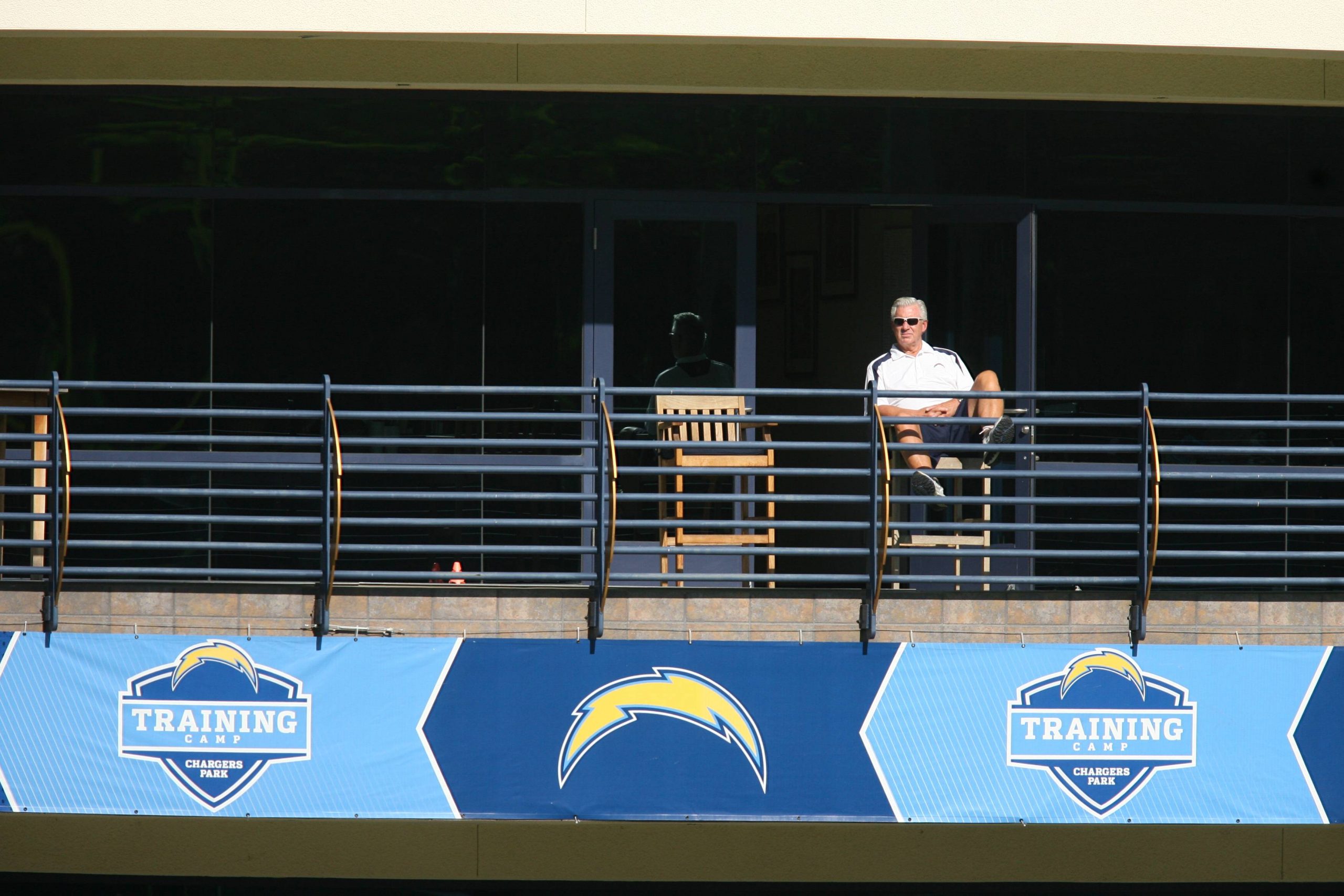 Aufzug Chargers - FootballR - NFL A.J. Smith - Ein Mann mit Sonnenbrille und weißem Poloshirt sitzt hinter einem Geländer mit dem Logo des NFL-Teams der Chargers. Er wirkt entspannt und beobachtet das Geschehen an einem sonnigen Tag von einem schattigen Balkon aus. Im Hintergrund mischen sich die General Manager verschiedener NFL-Teams untereinander. Diese Beschreibung wurde automatisch generiert.