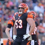 FootballR - NFL - Ein Footballspieler in der Uniform der Cincinnati Bengals steht mit den Händen in den Hüften auf dem Feld. Er trägt einen Helm mit orange-schwarzen Streifen, ein schwarzes Trikot mit der Nummer 53 und weiße Handschuhe und ähnelt damit dem ehemaligen NFL-Star Billy Price. Im Hintergrund sind andere Spieler und eine Menschenmenge im Stadion zu sehen. Diese Beschreibung wurde automatisch generiert.