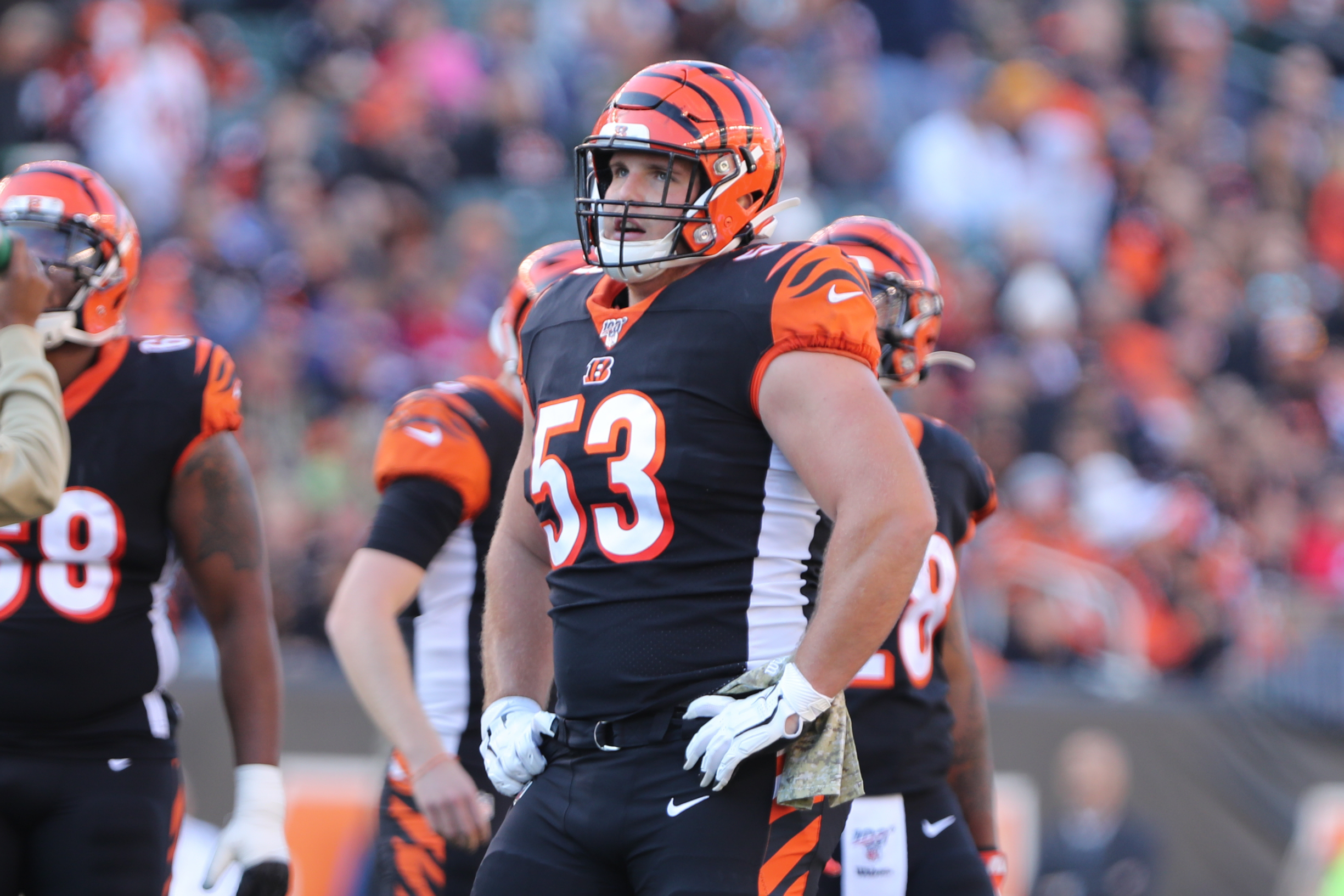FootballR - NFL - Ein Footballspieler in der Uniform der Cincinnati Bengals steht mit den Händen in den Hüften auf dem Feld. Er trägt einen Helm mit orange-schwarzen Streifen, ein schwarzes Trikot mit der Nummer 53 und weiße Handschuhe und ähnelt damit dem ehemaligen NFL-Star Billy Price. Im Hintergrund sind andere Spieler und eine Menschenmenge im Stadion zu sehen. Diese Beschreibung wurde automatisch generiert.