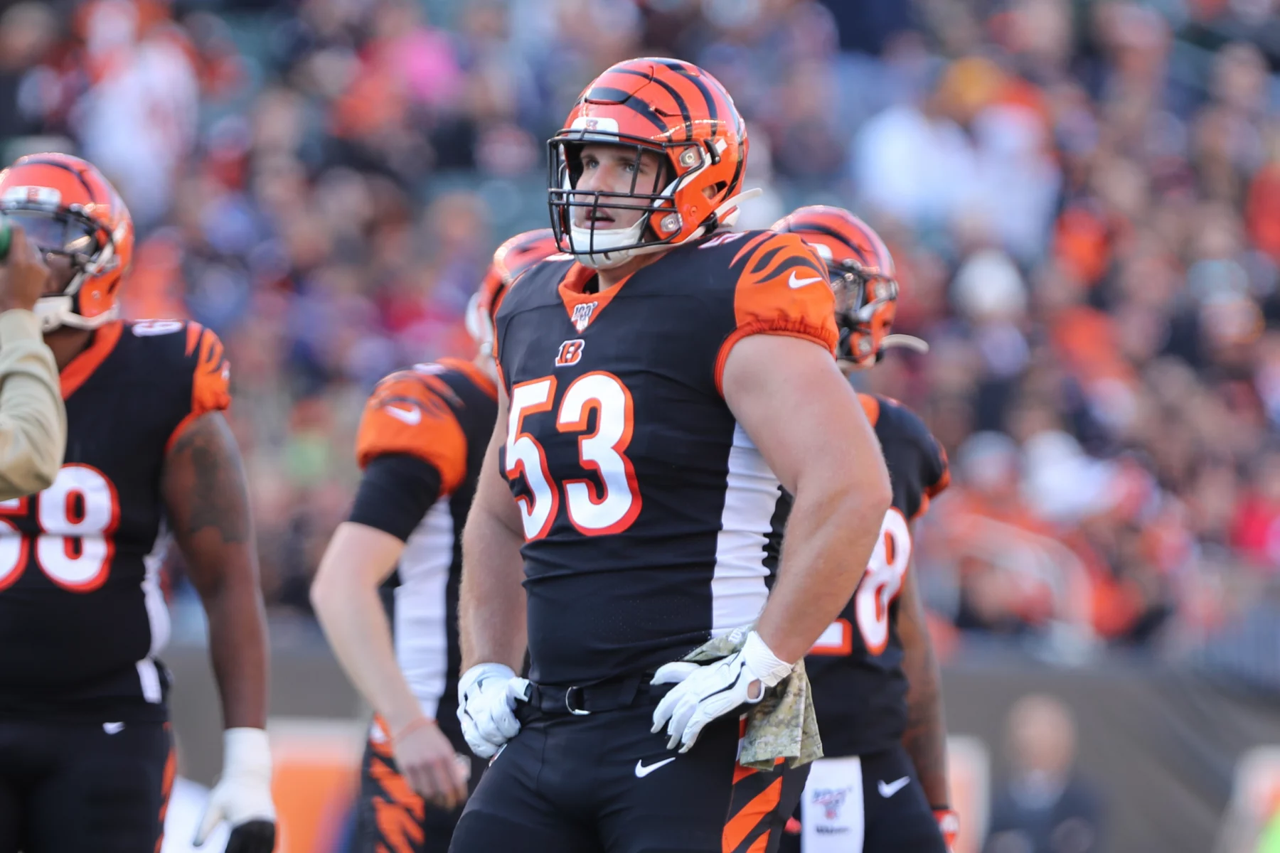 FootballR - NFL - Ein Footballspieler in der Uniform der Cincinnati Bengals steht mit den Händen in den Hüften auf dem Feld. Er trägt einen Helm mit orange-schwarzen Streifen, ein schwarzes Trikot mit der Nummer 53 und weiße Handschuhe und ähnelt damit dem ehemaligen NFL-Star Billy Price. Im Hintergrund sind andere Spieler und eine Menschenmenge im Stadion zu sehen. Diese Beschreibung wurde automatisch generiert.