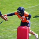 FootballR - NFL - Ein Footballspieler in einem orangefarbenen Broncos-Trikot mit der Nummer 41 und marineblauen Shorts übt während einer Trainingseinheit Handoffs. Der Spieler, der LB Drew Sanders ähnelt, trägt einen gepolsterten Helm und greift nach einem Football von einem Trainer in einem blauen Hemd, der einen angebundenen Ball hält. Im Vordergrund ist eine rote Tackling-Puppe zu sehen. Diese Beschreibung wurde automatisch generiert.