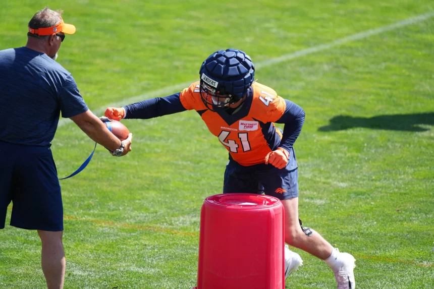 FootballR - NFL - Ein Footballspieler in einem orangefarbenen Broncos-Trikot mit der Nummer 41 und marineblauen Shorts übt während einer Trainingseinheit Handoffs. Der Spieler, der LB Drew Sanders ähnelt, trägt einen gepolsterten Helm und greift nach einem Football von einem Trainer in einem blauen Hemd, der einen angebundenen Ball hält. Im Vordergrund ist eine rote Tackling-Puppe zu sehen. Diese Beschreibung wurde automatisch generiert.
