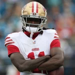 FootballR - NFL - Ein Footballspieler der San Francisco 49ers, in einem weißen Trikot mit roten Streifen und der Nummer 11, steht mit verschränkten Armen auf dem Feld. Der 49ers-Star Brandon Aiyuk, bekannt für seine beeindruckenden Leistungen und Dreadlocks, trägt einen goldenen Helm mit Gesichtsschutz. Im unscharfen Hintergrund sind andere Spieler und Zuschauer zu sehen. Diese Beschreibung wurde automatisch generiert.