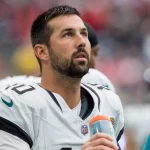 FootballR - NFL - Ein Footballspieler in weiß-schwarzer Uniform, der an Commanders Kicker Brandon McManus erinnert, blickt aufmerksam gen Himmel. Er hat kurze dunkle Haare, einen Bart und hält in seiner rechten Hand eine orangefarbene Wasserflasche. Im Hintergrund ist eine verschwommene Mischung aus Mitspielern und Zuschauern zu sehen, die für eine lebhafte Atmosphäre sorgt. Diese Beschreibung wurde automatisch generiert.