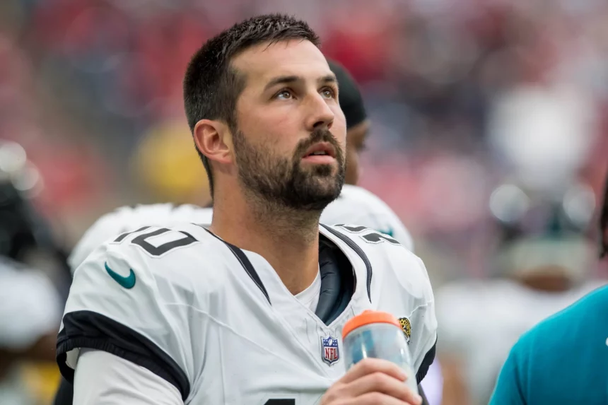 FootballR - NFL - Ein Footballspieler in weiß-schwarzer Uniform, der an Commanders Kicker Brandon McManus erinnert, blickt aufmerksam gen Himmel. Er hat kurze dunkle Haare, einen Bart und hält in seiner rechten Hand eine orangefarbene Wasserflasche. Im Hintergrund ist eine verschwommene Mischung aus Mitspielern und Zuschauern zu sehen, die für eine lebhafte Atmosphäre sorgt. Diese Beschreibung wurde automatisch generiert.