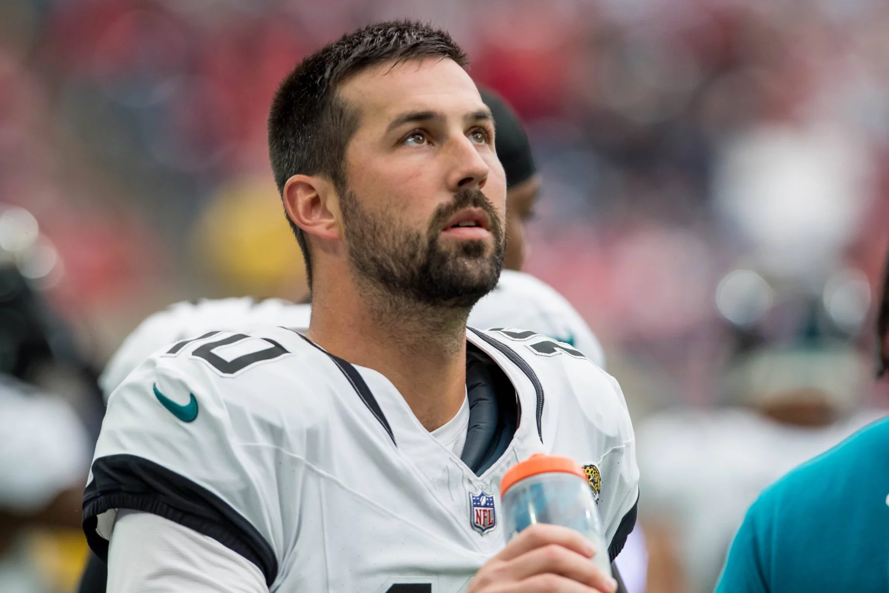 FootballR - NFL - Ein Footballspieler in weiß-schwarzer Uniform, der an Commanders Kicker Brandon McManus erinnert, blickt aufmerksam gen Himmel. Er hat kurze dunkle Haare, einen Bart und hält in seiner rechten Hand eine orangefarbene Wasserflasche. Im Hintergrund ist eine verschwommene Mischung aus Mitspielern und Zuschauern zu sehen, die für eine lebhafte Atmosphäre sorgt. Diese Beschreibung wurde automatisch generiert.