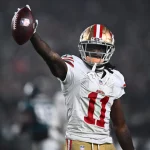 FootballR - NFL - Ein Footballspieler, der Brandon Aiyuk ähnelt, ein weißes San Francisco 49ers-Trikot mit der Nummer 11 und einen goldenen Helm mit roten Streifen trägt, hält einen Football in seiner rechten Hand. Im Hintergrund sind ein verschwommenes Stadion und andere Spieler zu sehen. Der Spieler wirkt triumphierend oder feierlich. Diese Beschreibung wurde automatisch generiert.