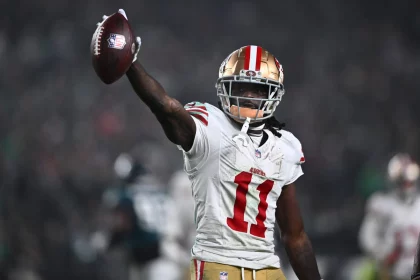 FootballR - NFL - Ein Footballspieler, der Brandon Aiyuk ähnelt, ein weißes San Francisco 49ers-Trikot mit der Nummer 11 und einen goldenen Helm mit roten Streifen trägt, hält einen Football in seiner rechten Hand. Im Hintergrund sind ein verschwommenes Stadion und andere Spieler zu sehen. Der Spieler wirkt triumphierend oder feierlich. Diese Beschreibung wurde automatisch generiert.