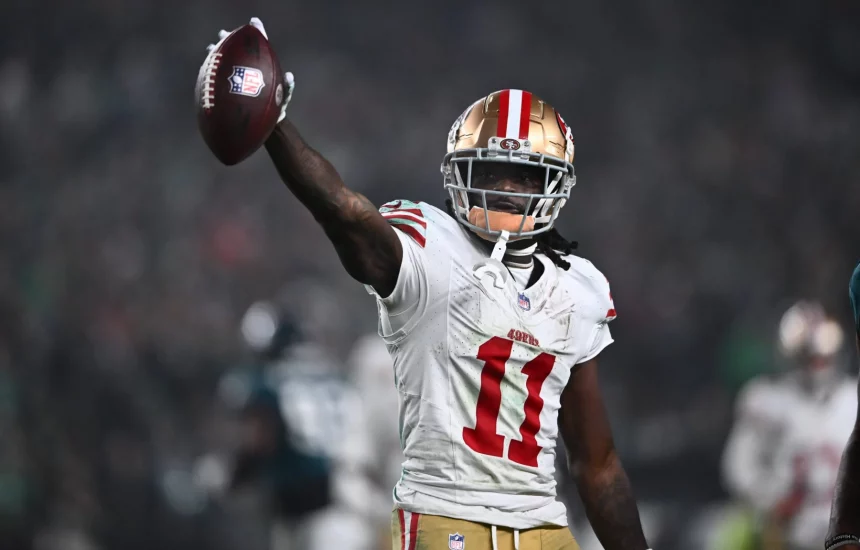 FootballR - NFL - Ein Footballspieler, der Brandon Aiyuk ähnelt, ein weißes San Francisco 49ers-Trikot mit der Nummer 11 und einen goldenen Helm mit roten Streifen trägt, hält einen Football in seiner rechten Hand. Im Hintergrund sind ein verschwommenes Stadion und andere Spieler zu sehen. Der Spieler wirkt triumphierend oder feierlich. Diese Beschreibung wurde automatisch generiert.