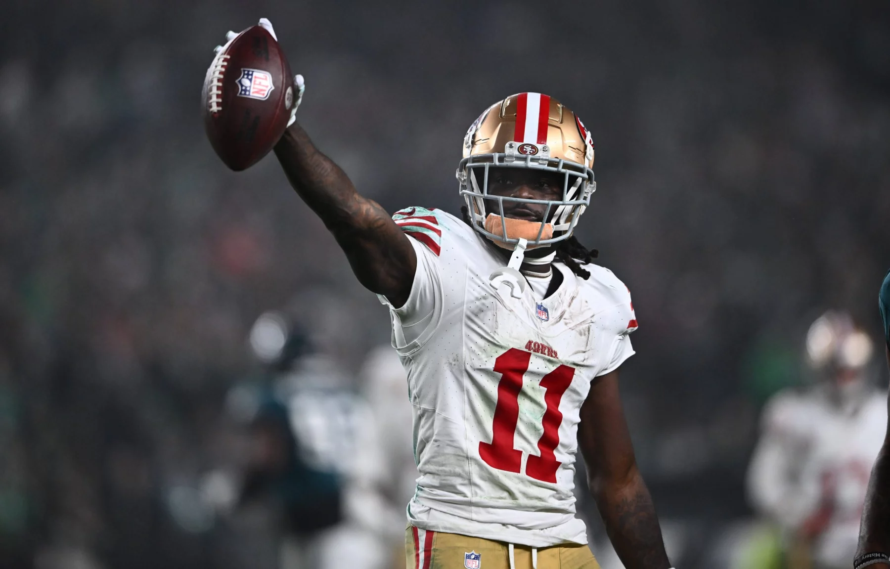 FootballR - NFL - Ein Footballspieler, der Brandon Aiyuk ähnelt, ein weißes San Francisco 49ers-Trikot mit der Nummer 11 und einen goldenen Helm mit roten Streifen trägt, hält einen Football in seiner rechten Hand. Im Hintergrund sind ein verschwommenes Stadion und andere Spieler zu sehen. Der Spieler wirkt triumphierend oder feierlich. Diese Beschreibung wurde automatisch generiert.