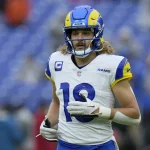 FootballR - NFL - Ben Skowronek - Ein professioneller Footballspieler, der eine blau-gelbe Uniform der Los Angeles Rams mit der Nummer 10 trägt, läuft über das Feld. Er hat blondes, lockiges Haar unter seinem blauen Helm mit einem Rams-Logo. Im Hintergrund ist ein großes, offenes Stadion mit blauen Sitzplätzen zu sehen. Diese Beschreibung wurde automatisch generiert.