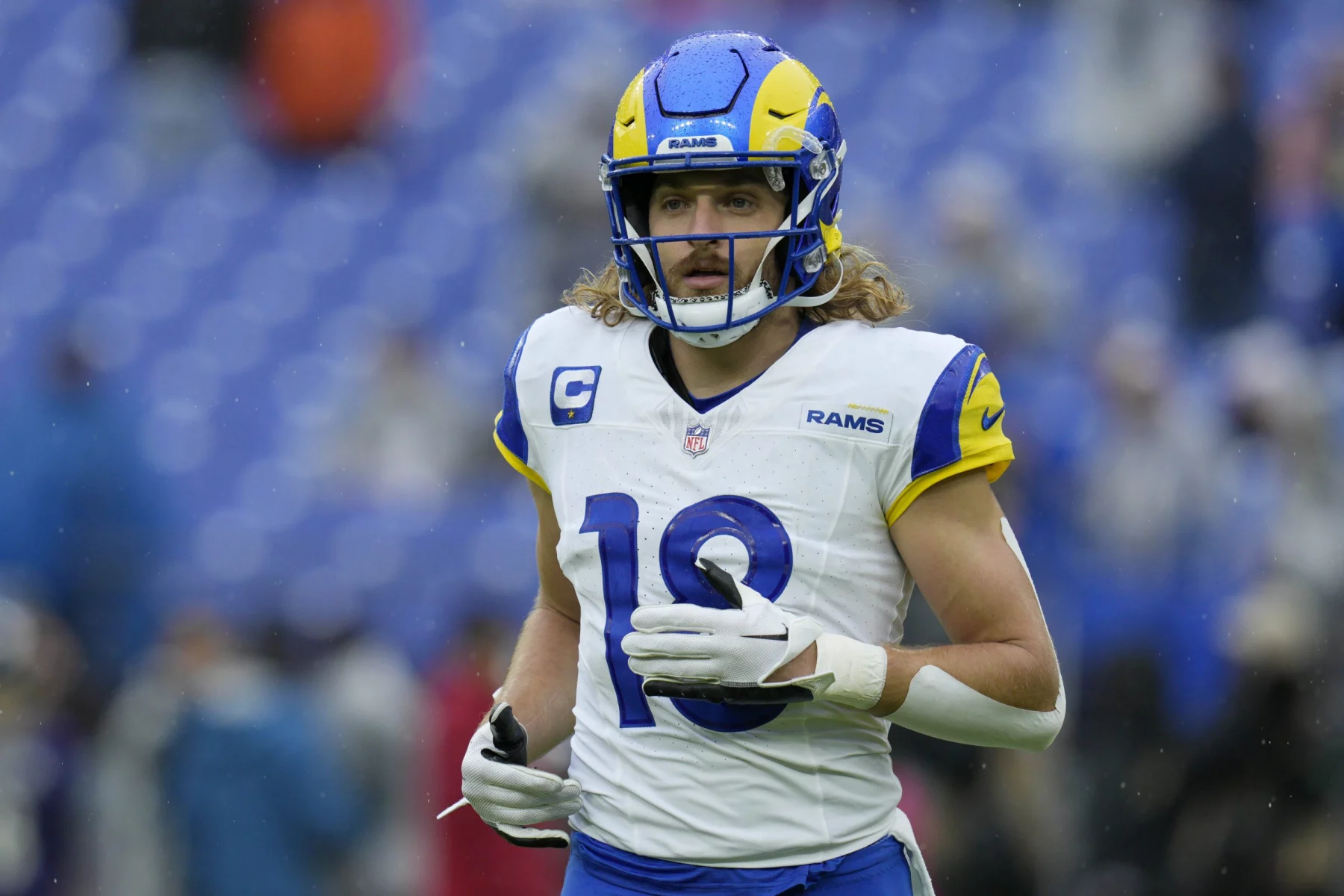 FootballR - NFL - Ben Skowronek - Ein professioneller Footballspieler, der eine blau-gelbe Uniform der Los Angeles Rams mit der Nummer 10 trägt, läuft über das Feld. Er hat blondes, lockiges Haar unter seinem blauen Helm mit einem Rams-Logo. Im Hintergrund ist ein großes, offenes Stadion mit blauen Sitzplätzen zu sehen. Diese Beschreibung wurde automatisch generiert.