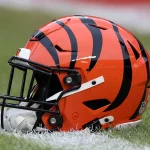 FootballR - NFL - Cincinnati Bengals - Ein orangefarbener Footballhelm mit schwarzen Tigerstreifen steht auf einem Rasenfeld im Paycor Stadium. Der Helm hat einen schwarzen Gesichtsschutz und einen weißen Kinnriemen, was das Team der Cincinnati Bengals symbolisiert. Im Hintergrund sind ein verschwommener Ausschnitt des Feldes und eine weiße Linie zu sehen. Diese Beschreibung wurde automatisch generiert.