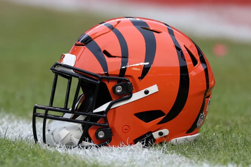 FootballR - NFL - Cincinnati Bengals - Ein orangefarbener Footballhelm mit schwarzen Tigerstreifen steht auf einem Rasenfeld im Paycor Stadium. Der Helm hat einen schwarzen Gesichtsschutz und einen weißen Kinnriemen, was das Team der Cincinnati Bengals symbolisiert. Im Hintergrund sind ein verschwommener Ausschnitt des Feldes und eine weiße Linie zu sehen. Diese Beschreibung wurde automatisch generiert.