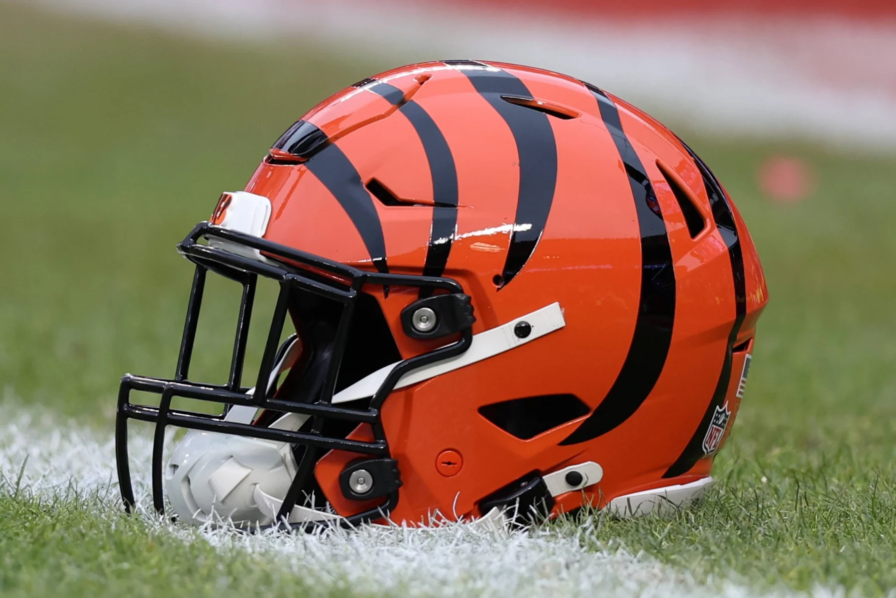 FootballR - NFL - Cincinnati Bengals - Ein orangefarbener Footballhelm mit schwarzen Tigerstreifen steht auf einem Rasenfeld im Paycor Stadium. Der Helm hat einen schwarzen Gesichtsschutz und einen weißen Kinnriemen, was das Team der Cincinnati Bengals symbolisiert. Im Hintergrund sind ein verschwommener Ausschnitt des Feldes und eine weiße Linie zu sehen. Diese Beschreibung wurde automatisch generiert.